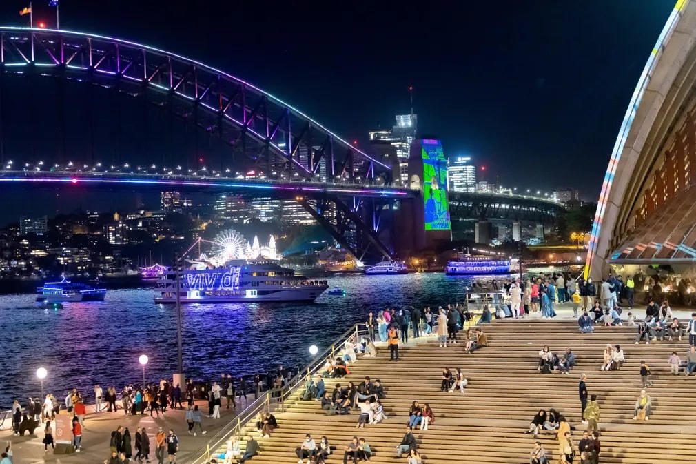 Người dân ngồi và đi lại tại tiền sảnh của Nhà hát Opera Sydney khi ánh đèn chiếu sáng một con thuyền và Cầu cảng Sydney để khai mạc lễ hội Vivid. Ảnh: James D Morgan / Getty Images.