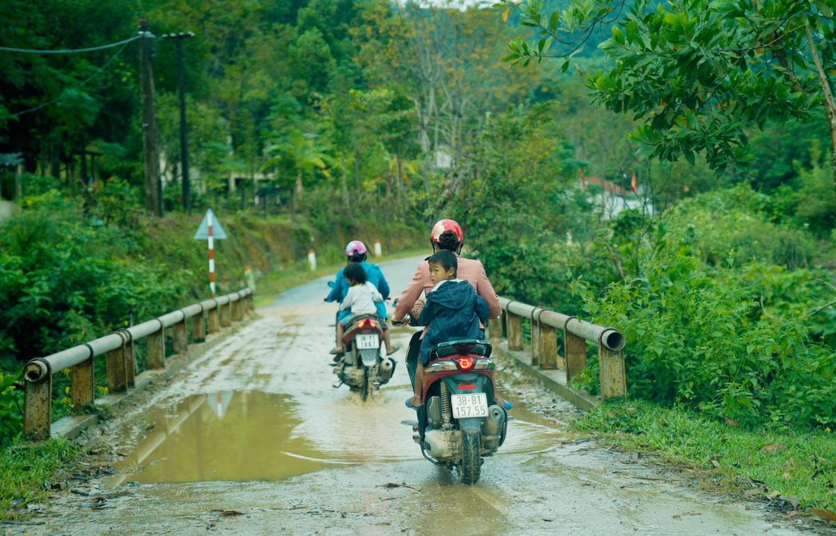 Trên chiếc xe máy cũ kỹ, men theo con đường bê tông ướt nhoẹt lấm đầy bùn đất, hai cô giáo vượt quãng đường hơn 3km mới đến được nhà của học sinh ở bản mới - khu tái định cư bản Rào Tre.