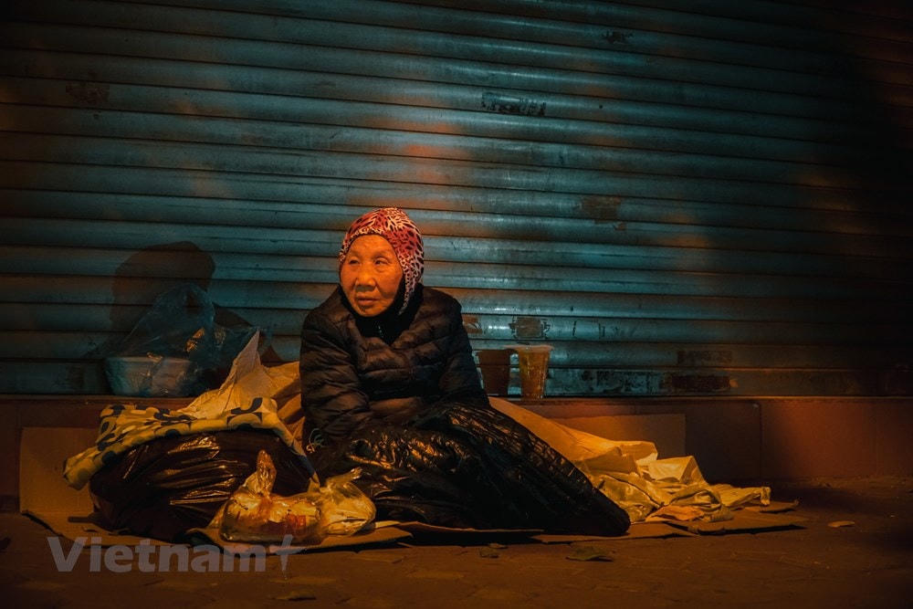 Ở vỉa hè góc phố Hai Bà Trưng giao với Quang Trung (Hà Nội), nhiệt độ thấp xuống kèm mưa lạnh khiến bà Nguyễn Thị Thanh khó vào giấc ngủ hơn mọi ngày. Theo lời bà Thanh, đây là nơi ở hằng ngày của bà để chờ đợi gom đủ tiền chữa trị cho đôi chân bị thương tật suốt 40 năm nay của mình.“Miếng cơm, manh áo cũng đều là từ những đơn vị từ thiện cung cấp cho tôi mỗi ngày. Tôi vẫn đang ngồi đợi người bạn sớm mang bảo hiểm y tế đến để được đi khám bệnh,” bà Thanh cho biết. (Ảnh: Minh Hiếu/Vietnam+).