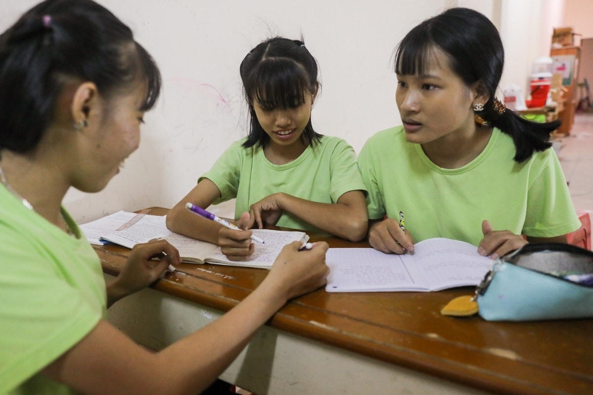 Ba chị em Yến Nhi (16 tuổi, góc phải) chăm chú trao đổi sau khi được giao bài tập. Cả ba chị em đều học hành dang dở, đang theo học chương trình lớp 5, ban ngày đi phụ bán quán phở. Ngoài ra, em trai của Nhi cũng đang học lớp 3 tại đây.  