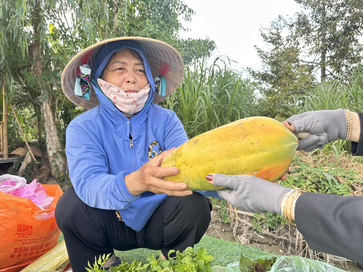 Bà Lê Thị Trúc Mai giới thiệu quả đu đủ “cây nhà lá vườn” do gia đình tự trồng với nụ cười hiền hậu, thân thiện, mến khách. (Ảnh: Trung Ngãi).