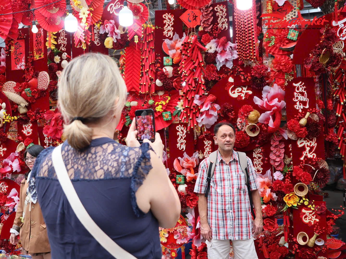 Du khách nước ngoài thích thú chụp ảnh lưu niệm bên gian hàng bán đồ trang trí Tết rực rỡ sắc màu tại Chợ hoa Xuân Phố cổ Hà Nội, ghi lại những khoảnh khắc ấn tượng và trải nghiệm không khí Tết cổ truyền Việt Nam..
